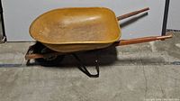 Side view of yellow metal wheelbarrow showing wooden handles and black metal wheel base.