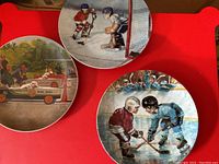 Three collectible plates displayed on a red surface showing vintage sports and car racing themes.