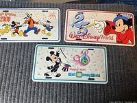 Three sealed Disney license plates laid on carpet, depicting colorful character and anniversary designs.