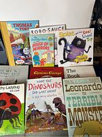 Photo shows a spread of 10 children's books arranged on a surface, titles visible include 'Thomas and Toby', 'Robo Sauce', 'The Berenstain Bears and the House of Mirrors', 'Splat and the Cool School Trip', 'Curious George', 'The Book with no Pictures', 'What the Dinosaurs Did Last Night', 'Leonardo the Terrible Monster', and 'Ladybug' themed book.