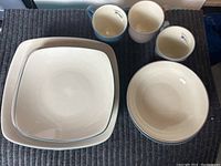 Three square plates stacked, two round bowls stacked, and three mugs (two beige, one blue)