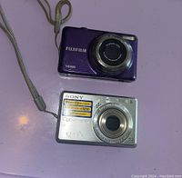 Top view of purple Fujifilm camera and silver Sony Cyber-shot 7.2MP camera on purple surface showing front lenses and bodies.
