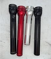 Four Maglite flashlights in black (2), red, and silver colors shown upright on a surface.
