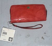 Closed red coral Anna Klein wallet showing textured snakeskin pattern, gold-tone logo plate, and attached wrist strap with price tag $48.