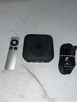 Apple TV device, remote control, and power cable on white surface showing front and side of device with remote and cable laid out
