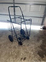 Photo showing a black metal wire basket on wheels with a tall handle, positioned indoors on a concrete floor with a garage door in the background.