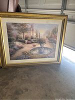 Photo of large framed wall picture showing a garden scene with a central water fountain, flowers, trees, and buildings in background, set against a gold ornate frame.