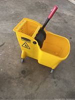 Photo of the yellow commercial mop bucket with red-handle wringer and caster wheels on a concrete floor.