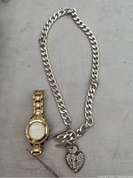 Photo showing gold-tone wristwatch and silver chunky chain necklace with heart pendant placed on grey surface