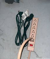 Photo showing three extension cords: one beige 5-outlet power strip with red on/off switch and two black cords with three-prong plugs, arranged on a light gray surface.