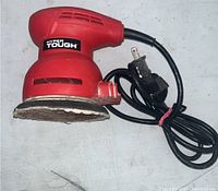 Red Hyper Tough electric orbital sander from side view showing body, plugged power cord, and worn sanding pad