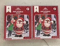 Two boxes featuring a 4 feet tall inflatable Santa decoration each, showing a child beside the inflated Santa for size comparison. Both boxed inflatables are new condition and priced $5 each.