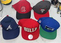 Five snapback hats arranged on a table showing team logos, indicating lot contents and wearable condition.