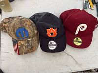 Three baseball caps on a white surface: camo Mopar logo cap with American flag tag, black Auburn Tigers cap, red Philadelphia Phillies New Era cap.
