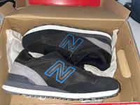 Side view of the pair of New Balance classic sneakers showing black upper with blue 'N' logo and grey suede accents inside the original box