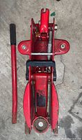 Top-down view of small red hydraulic car jack and its separate handle lying on a concrete surface showing some wear and use marks.