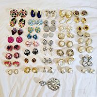 Photo showing 40 sets of vintage clip on earrings laid out on white fabric background. Variety of colorful, floral, pearl, and rhinestone designs.