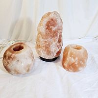 Three Himalayan salt pieces on white cloth: one lamp in center on wooden base, two candle holders on either side.