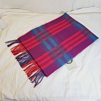 Folded purple, pink, red, and blue plaid wool scarf with fringed ends displayed flat on white surface