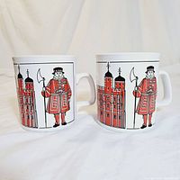 Front view of two white ceramic mugs featuring red Beefeater guards and Towers of London graphics side by side, showing detailed design and colors.