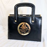 Front view of vintage black handbag showing gold-tone Asian character medallion, double top handles with stitched detail and gold hardware on white backdrop.