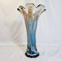 Tall handblown art glass vase shown against a white backdrop highlighting its swirl colors of blue, orange, and white with a scalloped flared rim and black base.