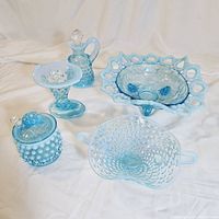 Eight light blue opalescent Fenton hobnail glass pieces arranged on white fabric background, showing candy dishes, honey mustard dish with lid and spoon, vinegar cruet, and cornucopia