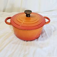 Front and lid view of Le Creuset Dutch oven showing flame orange enamel and wooden knob with some chips and wear on enamel.
