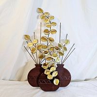 Front view of three wooden vases with geometric carved patterns mounting multiple metal stems with multi-colored brass petals in cascading arrangement.
