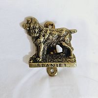 Front view of vintage metal door knocker depicting a standing Spaniel dog marked with 'SPANIEL' on the base, showing patina and age wear.