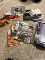 Lot of assorted automotive brochures and magazines featuring various car models including Dodge Challenger and branded magazines like TREAD, laid on carpet.