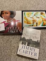Three books on a carpet: Michelle Obama photographic journey, Jimmy Carter's book, and a colorful abstract cover book.