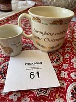 Photo showing Bunnykins Christening mug with decorative bunnies and text, alongside the smaller Peter Rabbit egg cup on a patterned tablecloth.