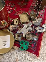 Assorted metal cookie cutters and green plastic cookie cutter placed on a carpet, alongside round tin container with a red floral design and gold lid.