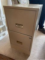 Front view of the beige metal filing cabinet with two drawers, showing brass recessed handles and lock on top drawer.
