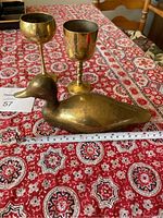 Photo showing one brass duck figurine and two brass goblets on a patterned red tablecloth, with a measuring tape indicating size approximately 5 inches tall for the duck.