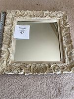 Front view of the square mirror showing the decorative carved frame in cream color and clear reflective glass surface.