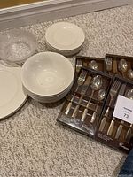Overview of assorted white plates and bowls next to four sets of boxed stainless steel cutlery.