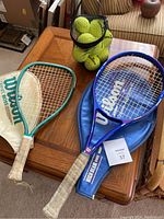 Two tennis racquets on a wooden table: a blue Wilson Graphite Aggressor, 27-inch tennis racquet with a white grip and blue cover, next to a smaller green tennis racquet approximately 19 inches long with white grip. A black mesh bag containing several tennis balls is also on the table.