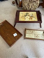 Three wooden serving trays; one with folding legs and floral design, one flat plain wood tray, and one with floral ceramic tile insets.