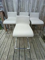 Front view of four modern bar chairs in a row, showing light beige seats and chromed metal legs.