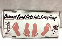 Front side of the metal sign showing "Damned Sand Gets Into Everything!" text with red footprints and beach illustrations
