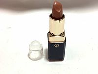 Top-down view of lipstick with transparent plastic lid beside it, showing the creamy beige shade with glossy finish