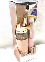 Front view of product packaging showing the foundation bottle with pink textured body and rose gold pump with bow, alongside a makeup brush.