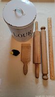 Full view of the vintage kitchenalia including the white metal flour tin, two wooden rolling pins, wooden butter patty mold, black ceramic pie bird, and wooden ruler for scale.