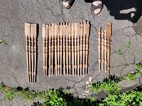 Fourteen antique wooden staircase spindles laid out on pavement; visible detailed turned design and measurement markings on some spindles; natural wood with signs of wear.