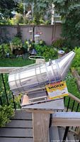 Photo of metal bee smoker on outdoor railing with garden background, showing full item and attached yellow label.
