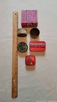 Top down photo showing all six tins including the pink box, ruler for scale, and assorted round and rectangular tins with various vintage labels.
