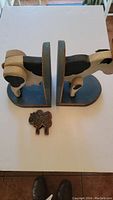 Pair of wooden cow bookends facing each other with a small wooden sheep figure between them on a white surface.
