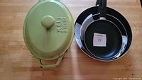 Set showing the green cast iron casserole with lid and three stacked black non-stick frying pans on a wooden surface.